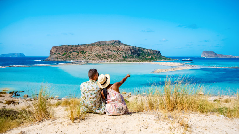 Blick auf die Lagune von Balos mit Sandbank und Insel; zwei Personen sitzen im Vordergrund auf einer Düne