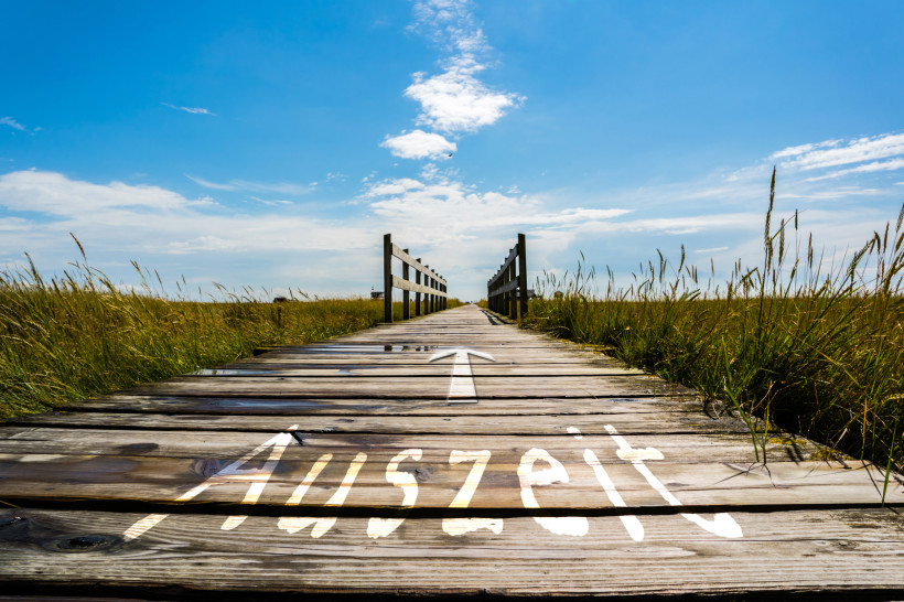 Holzsteg durch Dünen mit der Aufschrift Auszeit und Pfeil in Richtung Horizont