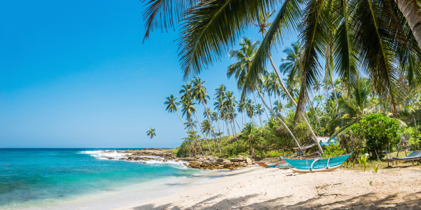 Panoramaaufnahme eines ruhigen tropischen Strandes mit weißem Sand, türkisblauem Meer und vielen Palmen. Im Vordergrund hängt ein traditionelles, blau gestrichenes Fischerboot zwischen zwei Palmen. Einige Wellen brechen leicht an den Felsen. Im Hintergrun