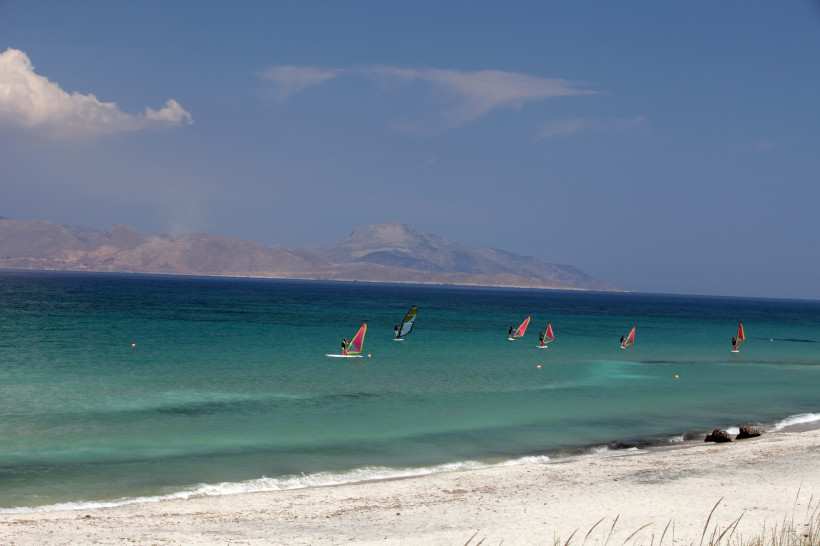 Kos - Mastichari  Mehrere Windsurfer gleiten auf türkisblauem Wasser vor der Küste von Kos. Der helle Sandstrand liegt ruhig im Vordergrund, dahinter ragen die Hügel und Berge einer benachbarten Insel am Horizont auf. Der Himmel ist klar mit wenigen Wolken