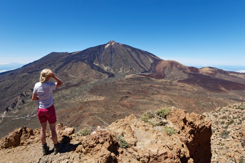 Eine Wanderin steht auf einem Felsen und schaut auf den Pico del Teide und die weite Vulkanlandschaft des Teide-Nationalparks auf Teneriffa.