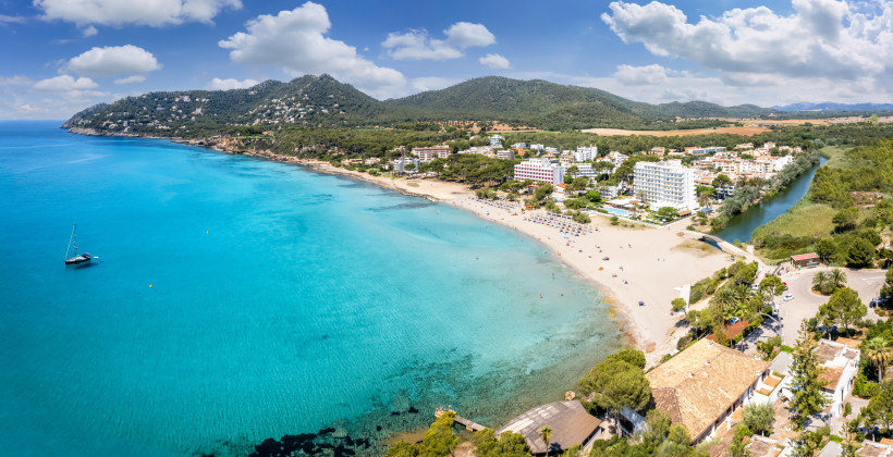 Luftaufnahme der Platja de Canyamel mit Sandstrand, türkisblauem Meer und Hotels vor bewaldeten Hügeln