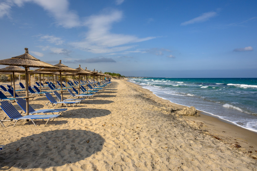 Kos Das Bild zeigt einen langen, sandigen Strand mit zwei Reihen leerer Liegestühle. Die Liegen sind blau gestreift und stehen unter großen Sonnenschirmen mit Strohdach. Die runden Schatten der Schirme liegen auf dem Sand. Rechts im Bild trifft das türkisfarb