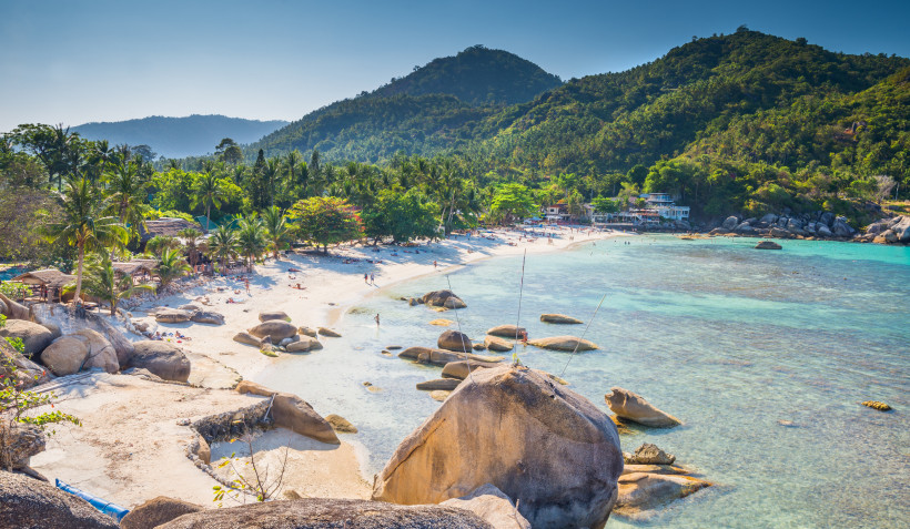 Thailand - Koh Samui Das Bild zeigt einen weitläufigen, tropischen Strand auf Koh Samui mit feinem, weißem Sand, umgeben von großen, runden Granitfelsen und Palmen. Das türkisfarbene, glasklare Meer zieht sich entlang der Küste, in der Ferne sind Menschen beim Schwimmen und S