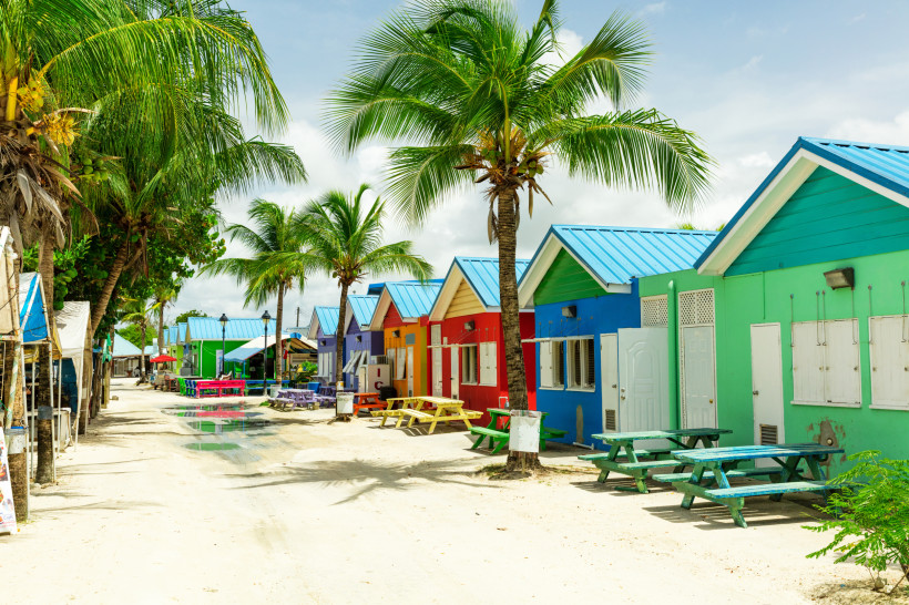Barbados Eine Straße auf Barbados mit bunten Strandhütten in kräftigen Farben wie Grün, Blau, Rot und Violett. Vor den Häusern stehen farblich passende Picknicktische unter Palmen. Die Straße ist sandig, mit einigen Pfützen, und wirkt ruhig und einladend.