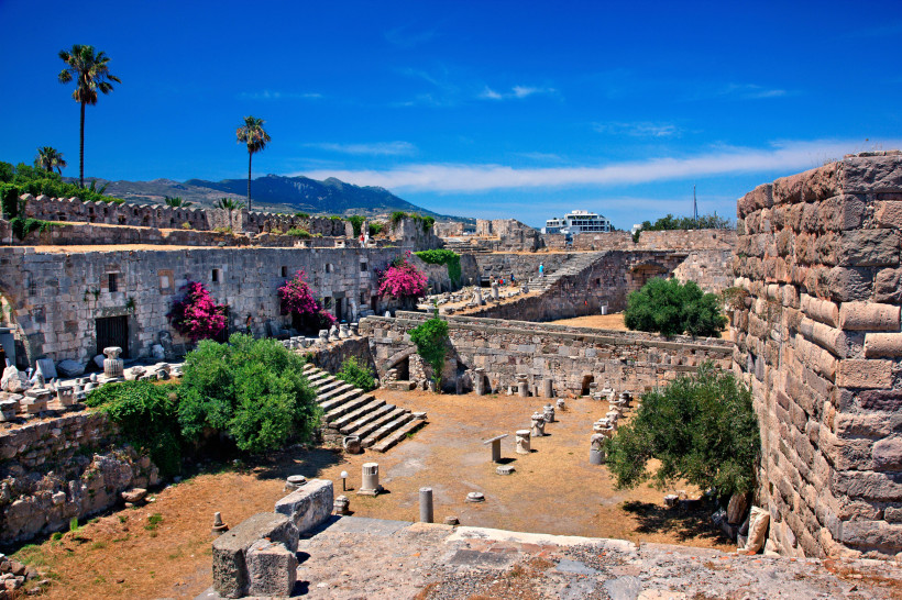 Kos - Kos-Stadt Mittelalterliche Festungsruinen auf Kos: massive Steinmauern, Stufen und verstreute Säulenfragmente, mit Bougainvillea und Palmen.