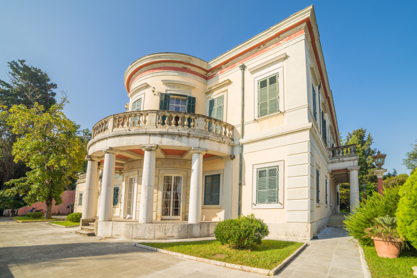 Eine elegante weiße Villa mit grünen Fensterläden, Rundbalkon und hohen Säulen im Eingangsbereich. Die Villa steht inmitten eines gepflegten Gartens mit Sträuchern und Bäumen, der Himmel ist klar und blau