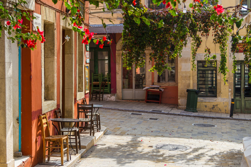 Straßenszene in Archanes mit Cafétischen und Stühlen vor farbigen Hausfassaden, überhängt von blühenden Bougainvilleen