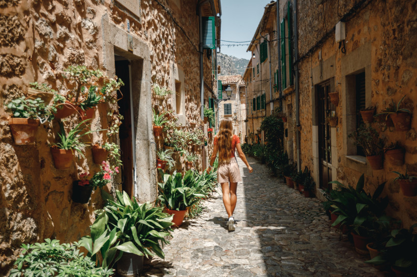 Schmale Kopfsteinpflastergasse in Palma de Mallorca mit Natursteinhäusern, Fensterläden und zahlreichen Pflanzen in Töpfen, Person geht entlang der Gasse