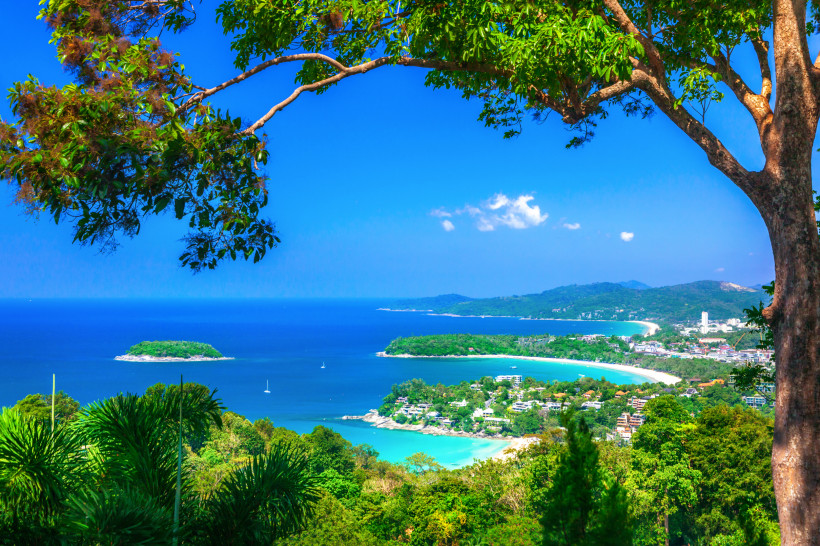 Thailand - Phuket Panoramablick über die Südwestküste der thailändischen Insel Phuket. Im Vordergrund rahmen dichte grüne Bäume das Bild ein, mit einem großen Ast im oberen Teil. Unten sieht man mehrere türkisfarbene Buchten mit weißen Sandstränden und einer vorgelagerten
