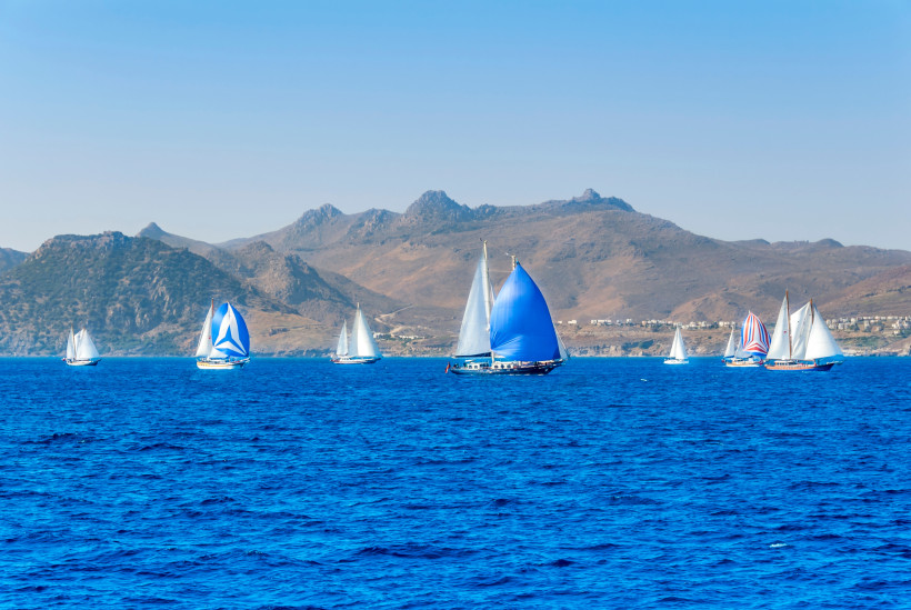 Mehrere Segelboote mit farbigen Segeln auf dem tiefblauen Wasser vor der bergigen Küste der türkischen Ägäis bei klarem Himmel