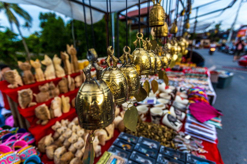 Thailand - Koh Samui Das Bild zeigt einen farbenfrohen Straßenmarkt in tropischer Umgebung, vermutlich auf Koh Samui. Im Vordergrund hängen goldene Metallglocken mit filigranen Gravuren und kleinen Blattanhängern. Auf den Tischen darunter liegen handgeschnitzte Holzfiguren in