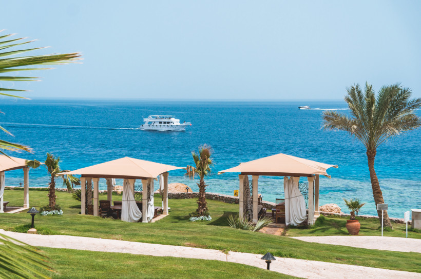 Ägypten - Makadi Bay Strandpavillons mit hellen Sonnendächern, Palmen und Blick auf das türkisfarbene Rote Meer in Makadi Bay, während Boote über das Wasser gleiten