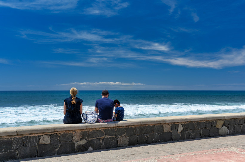 Drei Personen sitzen auf einer Ufermauer an der Strandpromenade von Maspalomas und blicken auf den Atlantik
