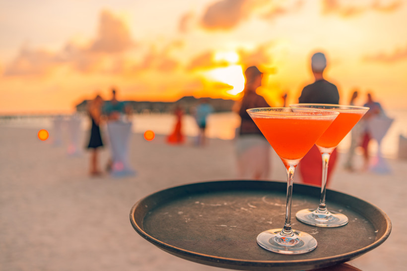 Zwei elegante Cocktailgläser auf einem Tablett mit verschwommener Strandparty und Sonnenuntergang im Hintergrund