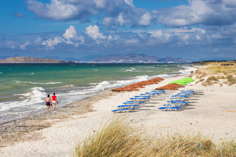 Kos - Mastichari  Zwei Personen spazieren am feinsandigen Strand entlang, das Meer brandet leicht ans Ufer. Rechts stehen Reihen von blauen Liegestühlen mit Sonnenschirmen, teils braun, teils grün. Im Hintergrund sind Hügel und Küstenlandschaft zu sehen, darüber ein dramat