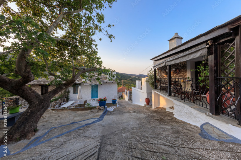 Traditioneller Dorfhof mit Steinhäusern, schattigem Baum, Terrasse und Blick auf die hügelige Landschaft auf der Insel Rhodos