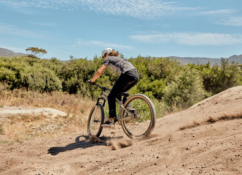 Südafrika Eine Person fährt mit einem Mountainbike über eine staubige Piste, umgeben von grüner Buschlandschaft und unter einem leicht bewölkten Himmel. Die Szene vermittelt sportliche Aktivität und Naturverbundenheit.