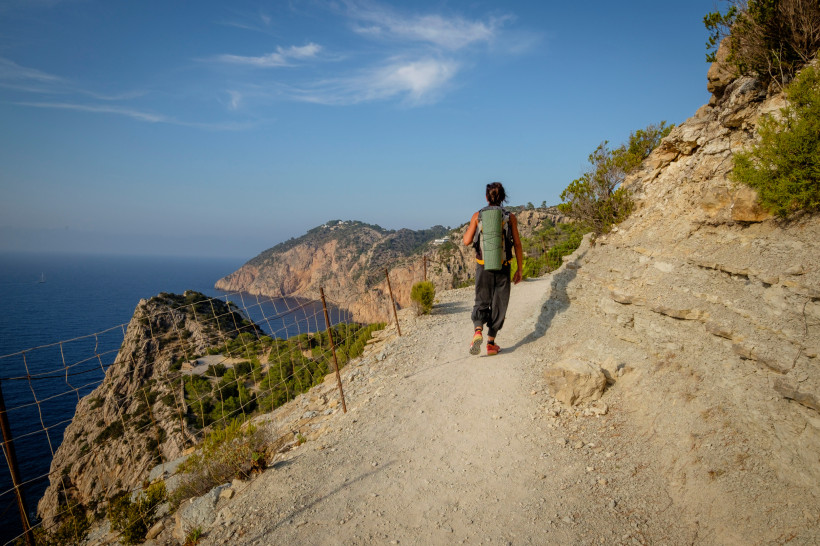 Ibiza Eine Person mit Rucksack wandert auf einem schmalen, steinigen Küstenpfad entlang einer steilen Klippe. Rechts türmen sich Felswände, links fällt der Blick weit über das tiefblaue Mittelmeer und grüne Küstenlandschaften. Der Himmel ist blau mit ein paar S