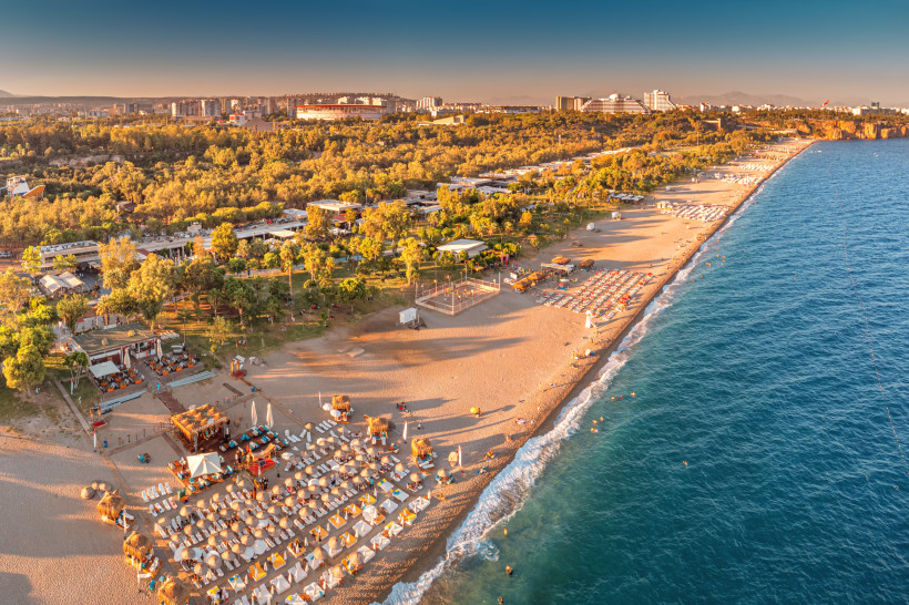 Konyaalti Beach Antalya – Sandstrand, Beachclubs und Promenade in der Abendsonne Luftaufnahme vom Konyaalti Beach in Antalya mit Strandliegen, Beachclubs, Promenade, grünen Parkanlagen und türkisblauem Meer bei Sonnenuntergang.