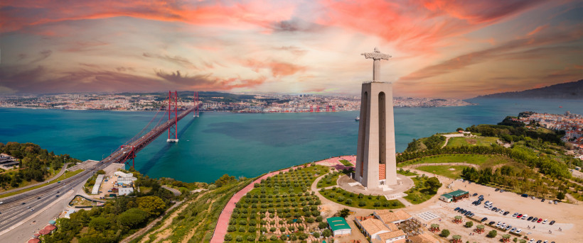 Lissabon Luftaufnahme der Christusstatue Cristo Rei in Lissabon mit ausgebreiteten Armen, die über dem Fluss Tejo thront. Links führt die rote Hängebrücke Ponte 25 de Abril über das türkisfarbene Wasser in die Stadt. Im Hintergrund erstreckt sich die Skyline von L