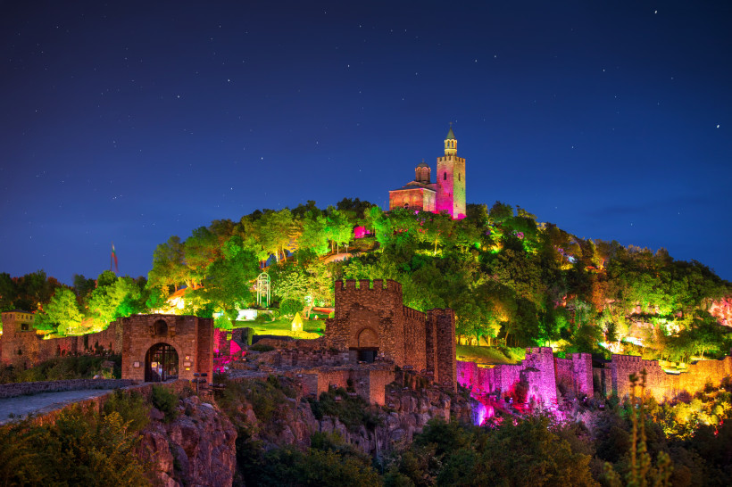 Festung Zarewez in Veliko Tarnovo, Bulgarien, bei Nacht. Die mittelalterliche Burganlage ist bunt beleuchtet – mit kräftigen Farben in Grün, Pink, Gelb und Violett. Im Hintergrund thront die Patriarchenkirche auf einem Hügel unter einem klaren Sternenhimm