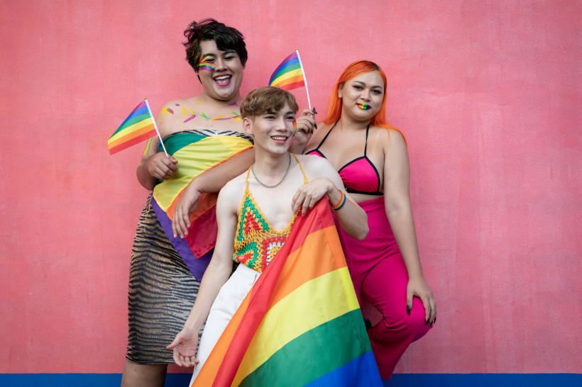 Thailand - Krabi Das Bild zeigt drei Personen, die selbstbewusst und fröhlich vor einer pinkfarbenen Wand posieren. Sie tragen farbenfrohe Kleidung in Pride-Farben, darunter ein Regenbogenhäkel-Top, ein pinkes Outfit und ein Wickeltuch mit Regenbogenflagge. Zwei der Perso
