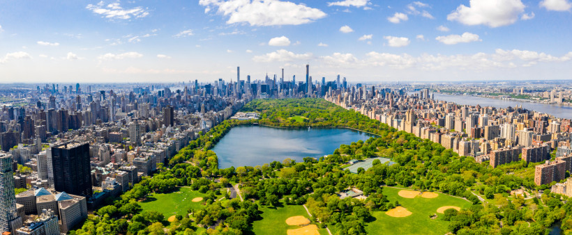 New York Luftaufnahme von New York City mit dem weitläufigen Central Park im Zentrum. Der Park ist von Hochhäusern und dicht bebauten Straßen eingerahmt. In der Ferne ist die Skyline von Manhattan mit zahlreichen Wolkenkratzern unter einem teils bewölkten blauen H