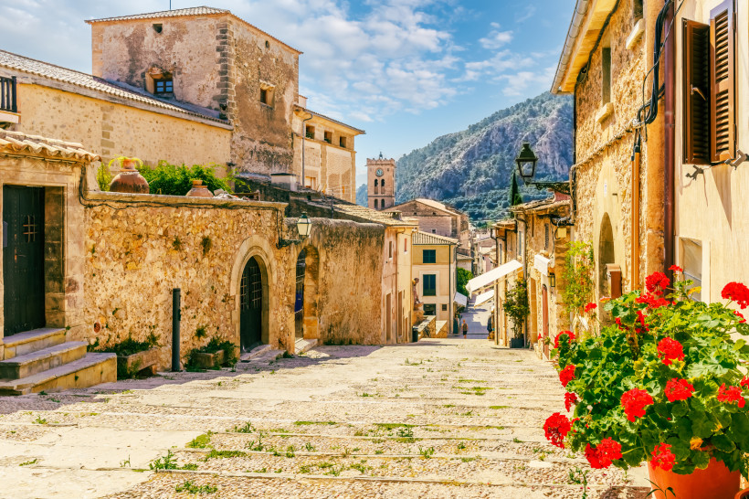 Mallorca - Pollença Historische Altstadtgasse von Pollença auf Mallorca. Die Calvari-Treppe zieht sich mit Kopfsteinpflasterstufen bergauf durch die Altstadt, flankiert von Natursteinhäusern mit Holztüren und Fensterläden. Links und rechts blühen rote Geranien in Töpfen. Im