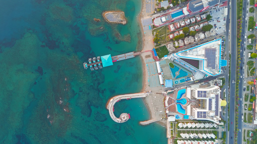 Senkrechte Drohnenaufnahme einer Hotelanlage in Türkler bei Alanya mit großem Poolbereich, direktem Strandzugang, Holzsteg mit Pavillons im Meer, Sonnenliegen am Strand und angrenzender Küstenstraße.