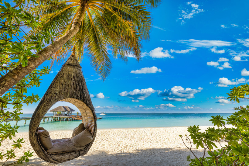 Strandpanorama, Mauritius Hängender Kokon-Sessel an einer Palme mit Blick auf weißen Sandstrand, türkisblaues Meer und Pavillon auf dem Wasser bei sonnigem Himmel