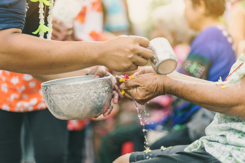 Eine jüngere Person gießt mit einer silbernen Schale Wasser über die Hände einer älteren Person. Blüten schwimmen im Wasser. Es ist eine traditionelle Geste des Respekts, vermutlich beim Songkran-Fest.