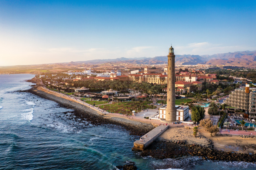 Luftaufnahme vom Leuchtturm von Maspalomas an der Küste Gran Canarias, flankiert von Promenade, Hotels und Bergen im Hintergrund.