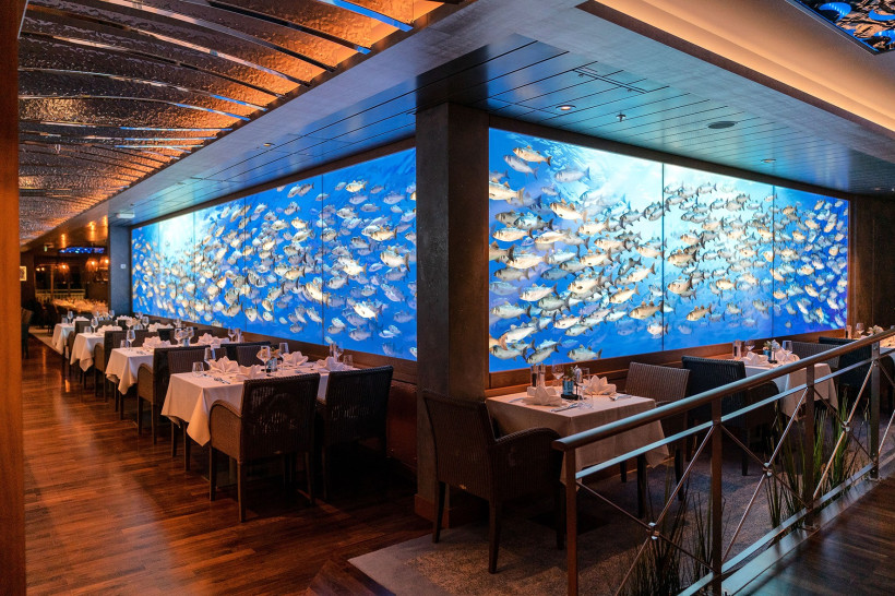 Fischrestaurant an Bord von AIDAnova mit gedeckten Tischen und Wandbild mit Fischschwarm Innenraum des Fischrestaurants an Bord von AIDAnova mit gedeckten Tischen und großer Wandprojektion eines Fischschwarms
