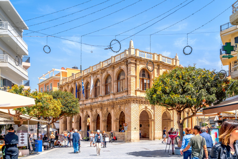 Platz in der Altstadt von Heraklion mit Arkadenbau, Bäumen und Passanten