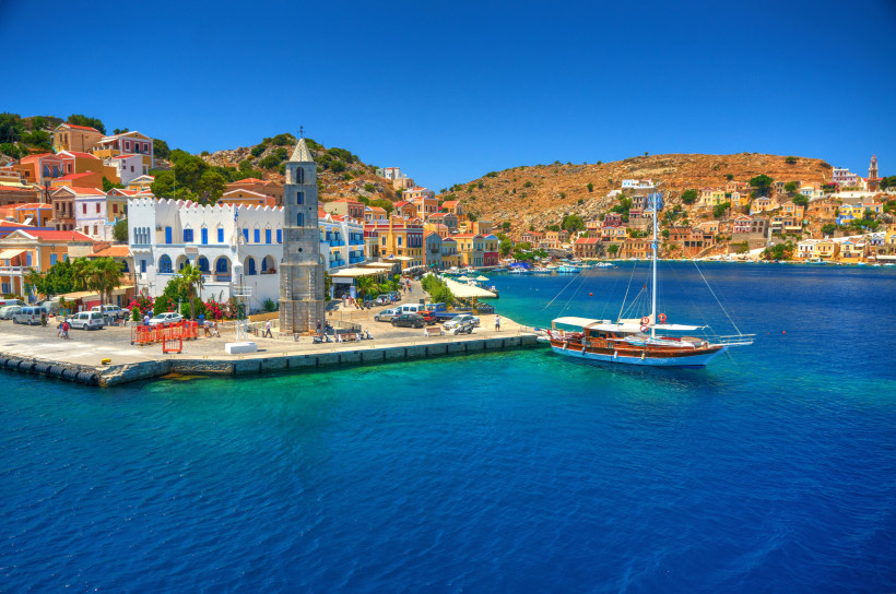 Blick auf den Hafen von Symi auf Rhodos mit bunten neoklassizistischen Häusern, Kirchturm und Segelboot im türkisblauen Meer