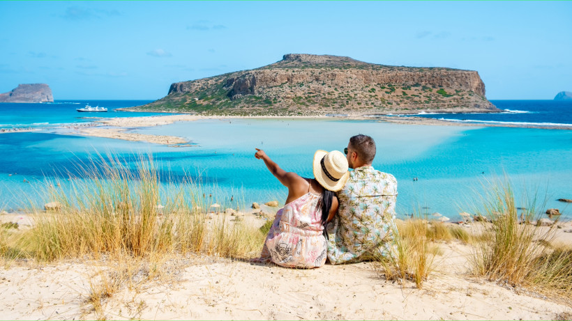 Kreta Ein Paar sitzt auf einer sandigen Düne mit Blick auf die türkisfarbene Lagune von Balos auf Kreta. Die Frau trägt einen Strohhut und zeigt mit dem Finger auf das Wasser, während der Mann neben ihr sitzt. Im Hintergrund liegen ein Boot, eine flache Landzun