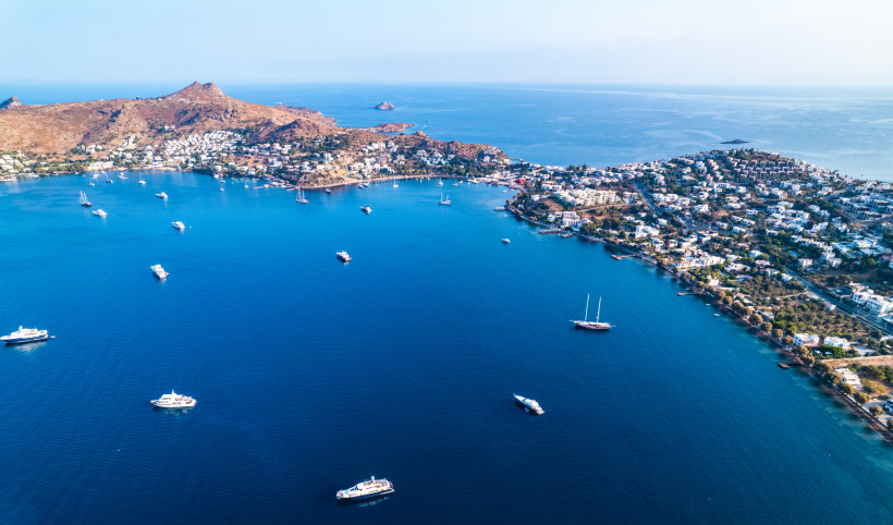 Luftaufnahme einer weitläufigen Bucht mit tiefblauem Meer, mehreren Yachten und einer dicht bebauten Küstenstadt an den Hängen