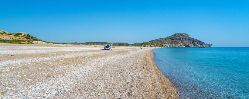 Weitläufiger Kiesstrand auf Rhodos mit ruhigem, klarem Meer, kaum Badegästen und hügeliger Landschaft im Hintergrund