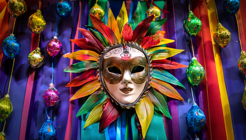 Detailaufnahme einer dekorativen venezianischen Karnevalsmaske mit goldenen Verzierungen und buntem Blattkranz, vor einem farbenfrohen Hintergrund mit glitzernden Ornamenten