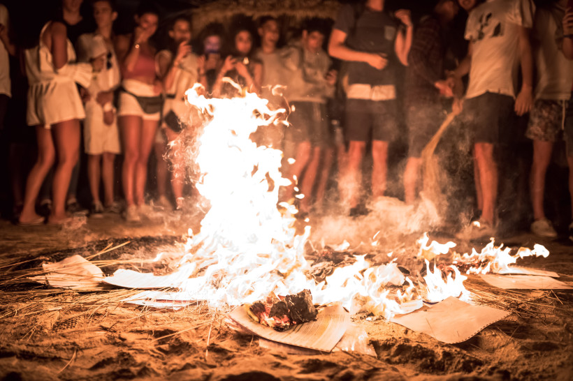 Ibiza Menschen stehen nachts barfuß um ein Feuer am Strand, das auf Holzstücken und Papier brennt. Der sandige Boden und der Rauch vermitteln eine warme, feierliche Atmosphäre. Einige Zuschauer filmen mit ihren Handys.