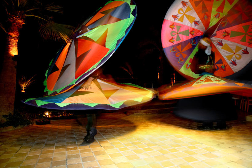 Ägypten Zwei Tänzer führen bei Nacht einen traditionellen Tanoura-Tanz mit leuchtend bunten, drehenden Kostümen auf. Die Szene spielt sich auf einem beleuchteten Platz mit Palmen ab.