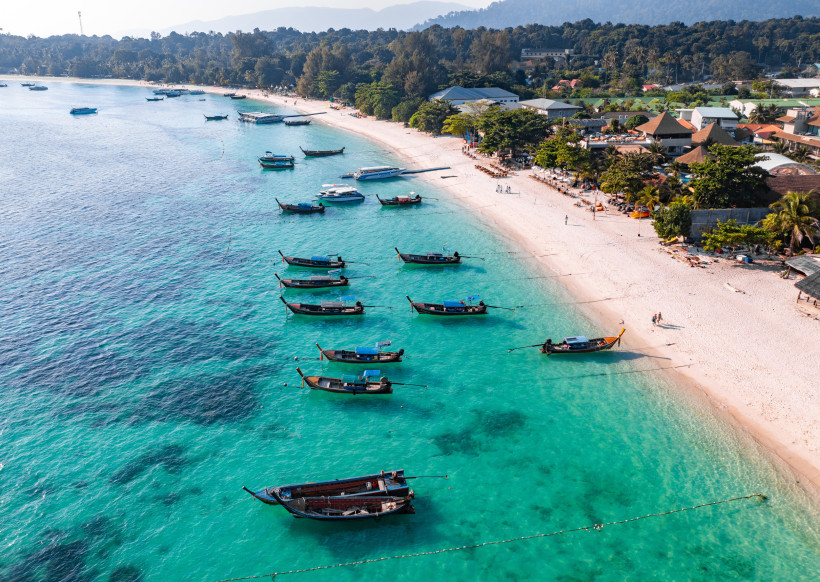 Thailand Luftaufnahme eines tropischen Strandes mit türkisblauem Wasser und traditionellen Longtail-Booten, die nahe der Küste ankern. Am weißen Sandstrand liegen gemütliche Resorts unter Palmen, im Hintergrund dichter Wald und Berge.