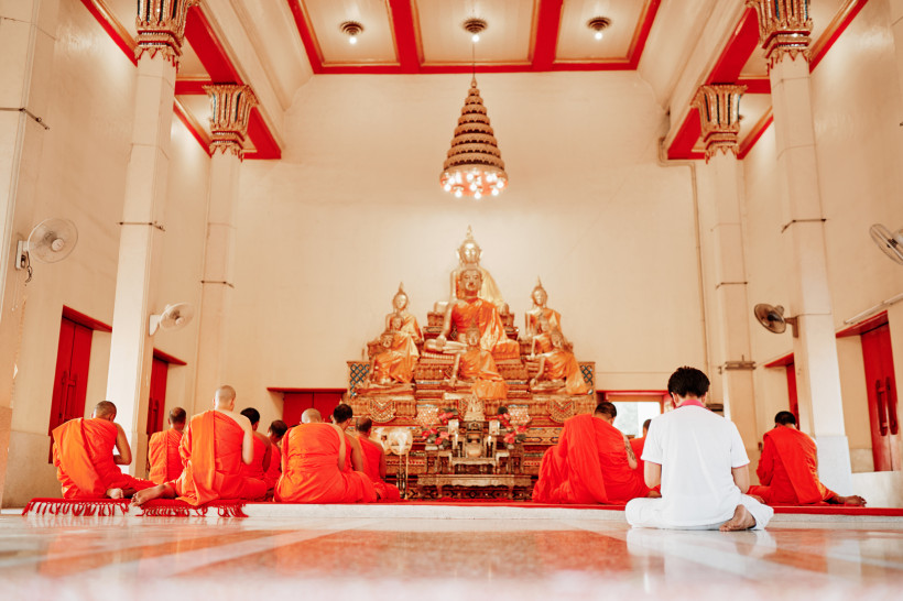 Thailand - Phuket Innenraum eines buddhistischen Tempels mit goldener Buddha-Statue. Eine Gruppe von Mönchen in orangenen Roben sitzt auf dem Boden in stiller Meditation, eine weitere Person in weißer Kleidung sitzt ihnen gegenüber. Die Atmosphäre wirkt friedlich und spiri
