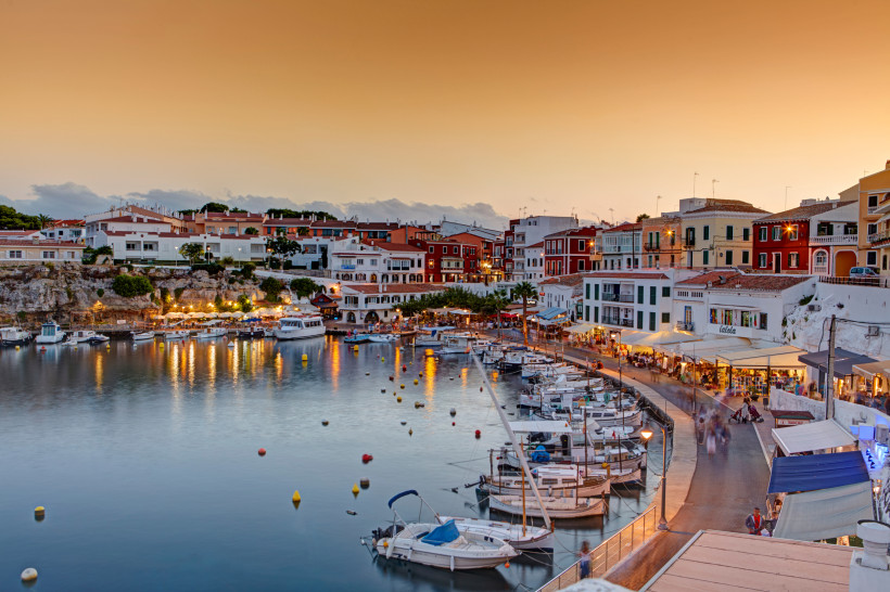 Hafenpromenade mit Booten und beleuchteten Häusern in Es Castell auf Menorca bei Abenddämmerung