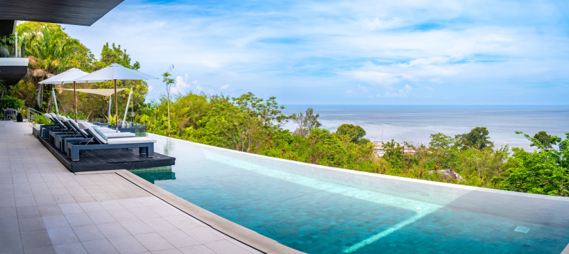 Thailand - Phuket Infinity-Pool einer modernen Unterkunft auf Phuket mit Blick aufs ruhige Meer. Neben dem Pool stehen bequeme Sonnenliegen mit weißen Sonnenschirmen. Dahinter tropische Vegetation und ein weiter, klarer Blick bis zum Horizont unter leicht bewölktem Himmel.