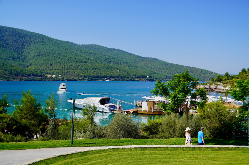 Blick auf eine ruhige Bucht bei Bodrum mit türkisfarbenem Meer, Booten, grünen Hügeln und Spazierweg am Ufer