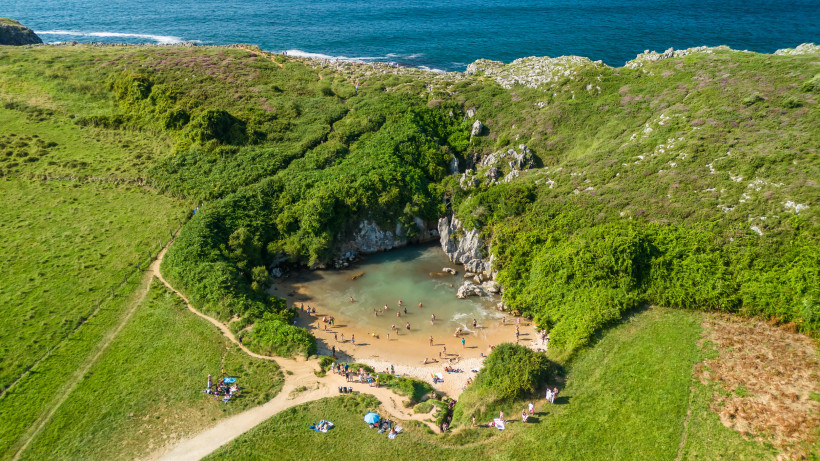 Luftaufnahme der Playa de Gulpiyuri mit kleiner Sandbucht, türkisfarbenem Wasser und grünen Hügeln an der Küste