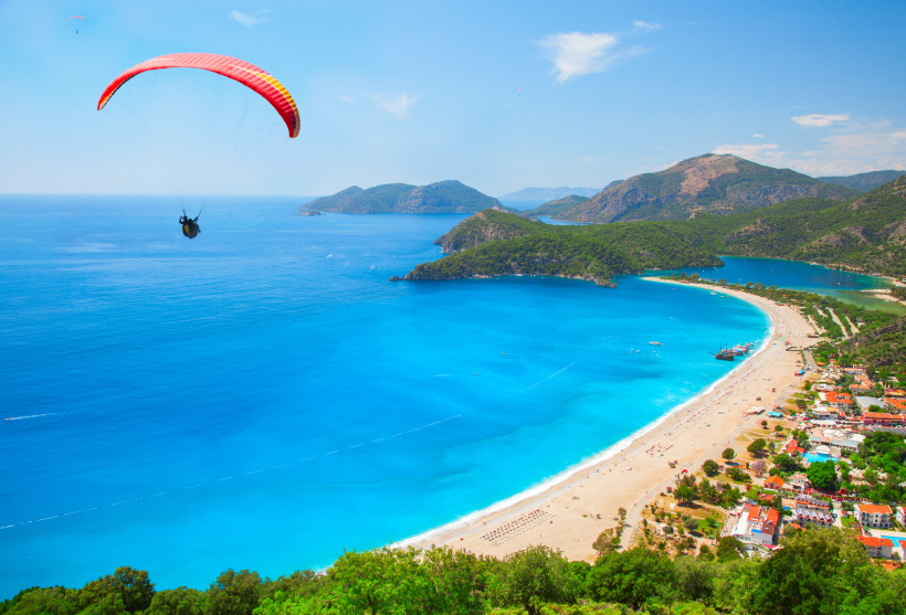 Paraglider über dem türkisblauen Meer und dem berühmten Strand von Ölüdeniz in der Türkei. Im Hintergrund eine traumhafte Küstenlandschaft mit grünen Hügeln und feinem Sandstrand