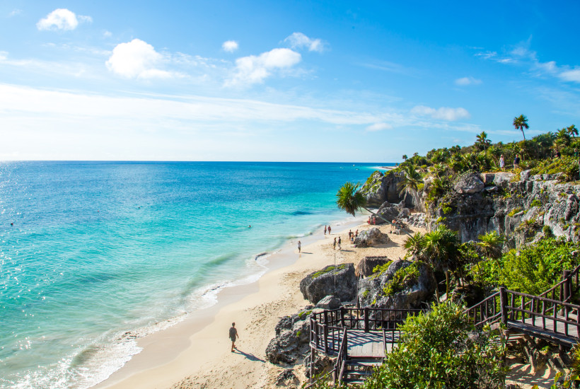 Winter in Mexiko Wunderschöner Sandstrand mit türkisblauem Meer, umgeben von Felsen und tropischer Vegetation. Menschen spazieren und entspannen am Strand unter einem sonnigen, blauen Himmel